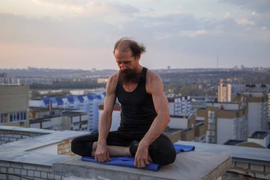 Orta yaşlı bir adam çatıda yoga yapıyor. Gün batımında çatıda yoga..