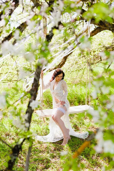 Jeune mariée séduisante en négligée blanche et lingerie assise sur une balançoire blanche — Photo