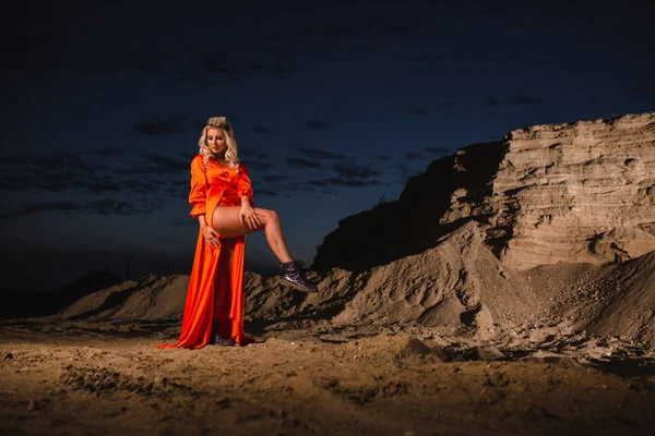 Kvinna i orange klänning poserar nära sand kulle i bakgrunden av solnedgången — Stockfoto