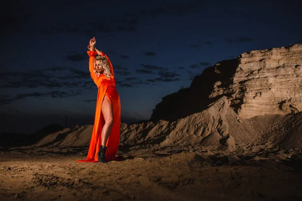 日没の背景に砂の丘の近くにポーズオレンジのドレスの女性 ストック写真