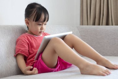 Asyalı Çinli küçük kız dijital tablet evde oturma odasında oynarken kanepede oturan