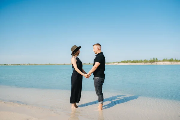 黒い服を着た女の子を持つ若いカップルが青い水の端の白い砂の上を歩いています — ストック写真