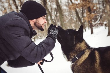 Ceketli ve örgü şapkalı bir adam Amerikan Akita köpeğiyle karlı bir ormanda yürüyor.