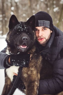 Ceketli ve örgü şapkalı bir adam Amerikan Akita köpeğiyle karlı bir ormanda yürüyor.