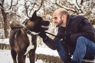 Ceketli ve örgü şapkalı bir adam Amerikan Akita köpeğiyle karlı bir ormanda yürüyor.