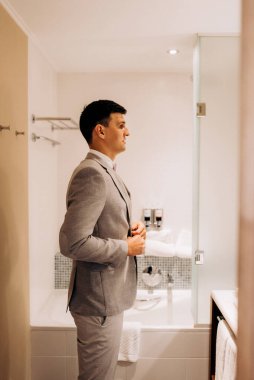 Üç parçalı takım elbiseli bir damat sabah bir otel banyosunda aynalı bir düğüne hazırlanır.