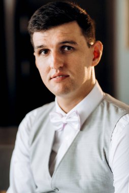 Otel lobisinde oturan takım elbiseli ve beyaz papyonlu bir damadın portresi.