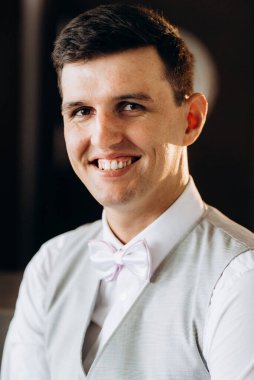 Otel lobisinde oturan takım elbiseli ve beyaz papyonlu bir damadın portresi.