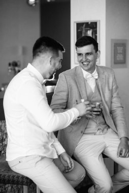 Bir otel lobisinde gelini bekleyen bir arkadaşla oturan takım elbiseli ve beyaz papyonlu bir damadın portresi.