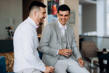 Bir otel lobisinde gelini bekleyen bir arkadaşla oturan takım elbiseli ve beyaz papyonlu bir damadın portresi.