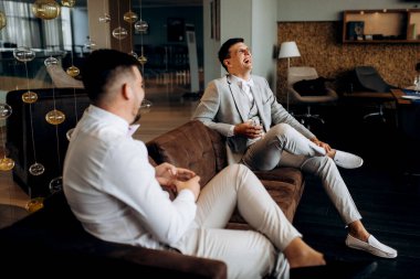 Bir otel lobisinde gelini bekleyen bir arkadaşla oturan takım elbiseli ve beyaz papyonlu bir damadın portresi.