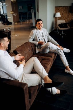 Bir otel lobisinde gelini bekleyen bir arkadaşla oturan takım elbiseli ve beyaz papyonlu bir damadın portresi.