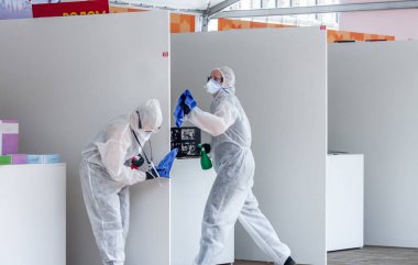 Beyaz koruyucu takım elbiseli, el dezenfektanı, temizlik süngeri ve tek kullanımlık eldivenlerle kamuya açık alanda yüzeyi silen iki kadın doktor. Dezenfeksiyon temizleme servisi virüs enfeksiyonlarını durdurdu..