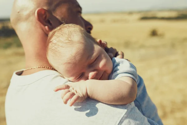 Der junge Vater hält ein neugeborenes Baby auf seiner Schulter, das in der Natur spazieren geht. Der glückliche Vater trägt kurze Hosen und ein T-Shirt. Internationaler Vatertag — Stockfoto