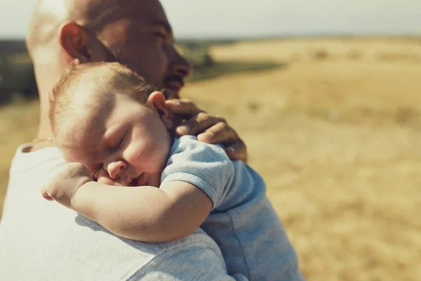 Der junge Vater hält ein neugeborenes Baby auf seiner Schulter, das in der Natur spazieren geht. Der glückliche Vater trägt kurze Hosen und ein T-Shirt. Internationaler Vatertag — Stockfoto