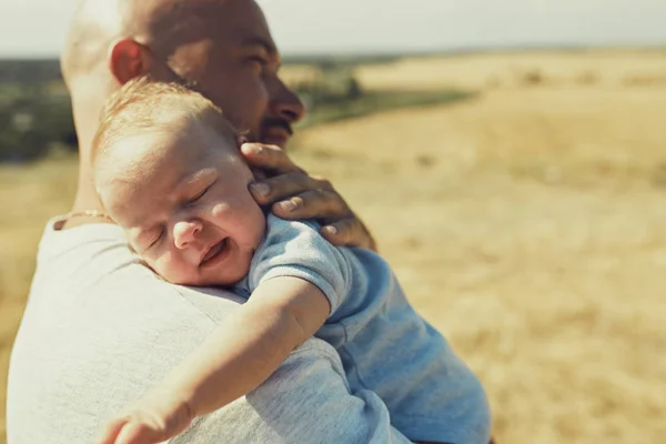 Der junge Vater hält ein neugeborenes Baby auf seiner Schulter, das in der Natur spazieren geht. Der glückliche Vater trägt kurze Hosen und ein T-Shirt. Internationaler Vatertag — Stockfoto