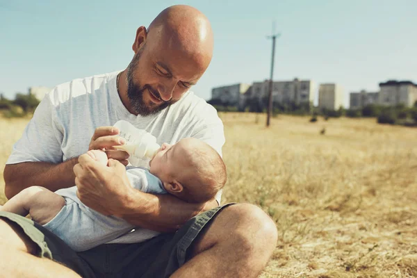 年轻的爸爸给一个刚出生的婴儿从瓶子里吃，一个假人坐在大自然的草地上。快乐的父亲穿着短裤和T恤。国际父亲节 — 图库照片