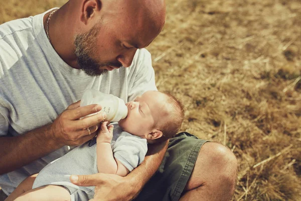 年轻的爸爸给一个刚出生的婴儿从瓶子里吃，一个假人坐在大自然的草地上。快乐的父亲穿着短裤和T恤。国际父亲节 — 图库照片