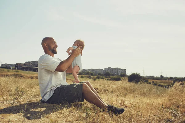 年轻的爸爸把一个刚出生的婴儿抱在伸出的胳膊上。快乐的父亲穿着短裤和T恤。国际父亲节 — 图库照片