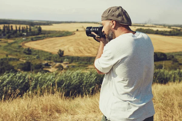 Bir kamera ile fotoğrafçı tahıl alanlarının arka planda duruyor. şort ve tişört giyen bir adam, kafasına bir şapka — Stok fotoğraf