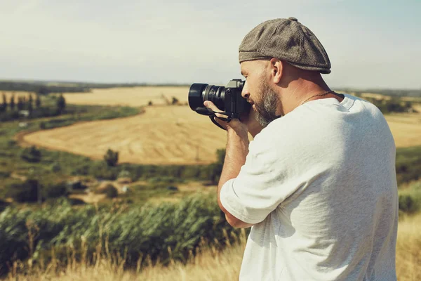 카메라를 들고 있는 사진작가가 시리얼 밭을 배경으로 서 있습니다. 반바지와 티셔츠를 입은 남자, 머리에 모자 — 스톡 사진