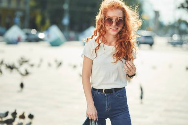 Beyaz tişört ve güneş gözlüğü ile güzel bir kızıl saçlı kız sokakta yürür ve çerçeve içinde gülümsüyor