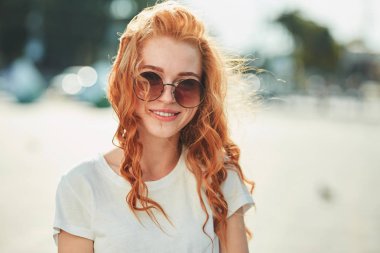 Beyaz tişört ve güneş gözlüğü ile güzel bir kızıl saçlı kız sokakta yürür ve çerçeve içinde gülümsüyor