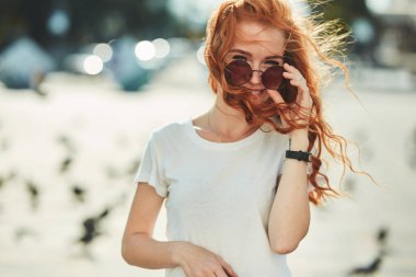 Güzel kızıl saçlı kız sokakta eğleniyor. Kızlar güzel bir rakam, beyaz bir t-shirt ve güneş gözlüğü ile kot var