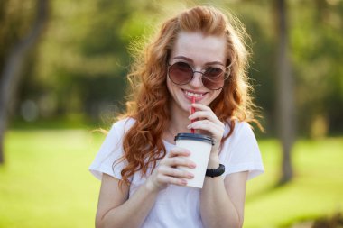 güzel kızıl saçlı kız bir fincan kahve ile parkta yürür. Kız kameraya gülümsüyor. Güneş gözlüklüğü olan bir kız ve kolunda modern bir saat.