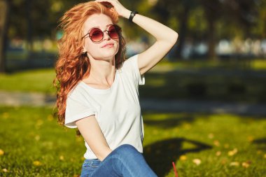 Güzel bir kızıl saçlı kız, güneş ışınları tarafından ısınmış, bir çim üzerinde oturuyor ve kamera için poz. Kız kot pantolon, yüzünde gözlük ve kolunda modern bir alet olan bir tişört giyiyor.