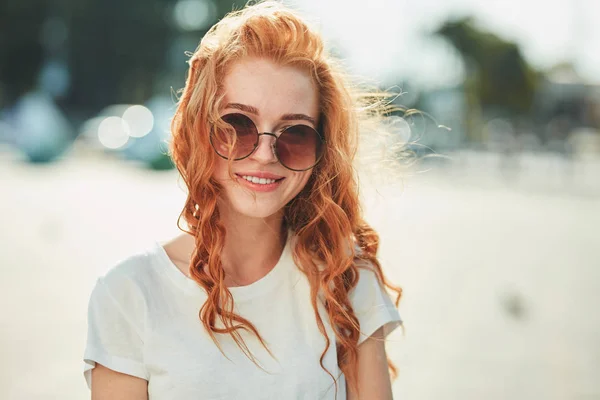 Una hermosa chica pelirroja con una camiseta blanca y gafas de sol camina por la calle y sonríe en el marco — Foto de Stock