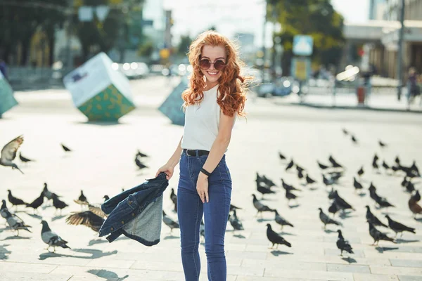 Krásná zrzavá dívka se na ulici baví. Děvčata mají krásnou postavu, bílé tričko a džíny s brýlemi — Stock fotografie