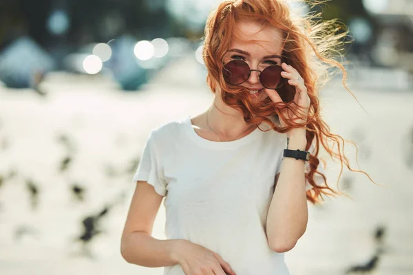 Vacker rödhårig flicka ha kul på gatan. Flickorna har en vacker figur, en vit T-shirt och jeans med solglasögon — Stockfoto