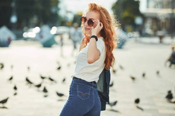 Hermosa chica pelirroja divirtiéndose en la calle. Las chicas tienen una hermosa figura, una camiseta blanca y jeans con gafas de sol —  Fotos de Stock