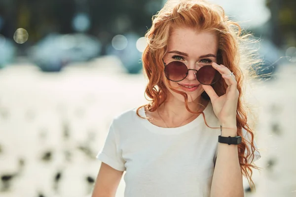 Güzel kızıl saçlı kız sokakta eğleniyor. Kızlar güzel bir rakam, beyaz bir t-shirt ve güneş gözlüğü ile kot var — Stok fotoğraf