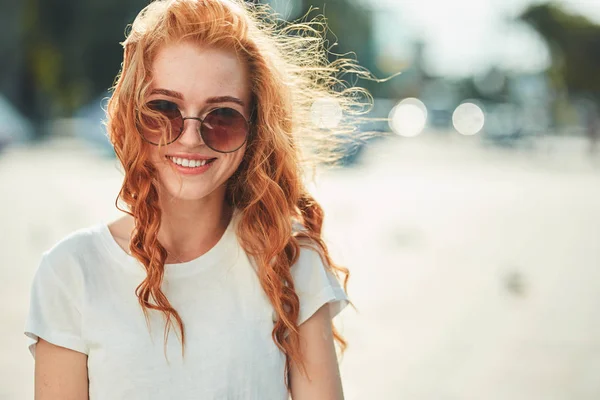 Una hermosa chica pelirroja con una camiseta blanca y gafas de sol camina por la calle y sonríe en el marco —  Fotos de Stock