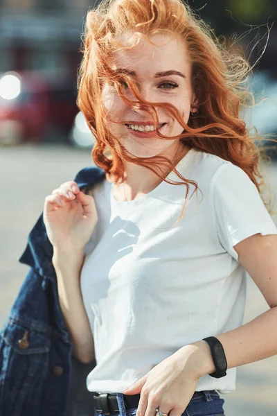 Vacker rödhårig flicka ha kul på gatan. Flickorna har en vacker figur, en vit T-shirt och jeans med solglasögon — Stockfoto
