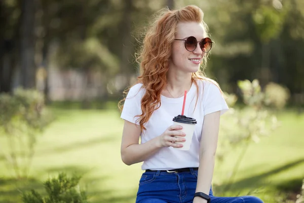 Hermosa chica pelirroja camina por el parque con una taza de café. La chica sonríe a la cámara. Una chica con gafas de sol y un reloj moderno en su brazo —  Fotos de Stock