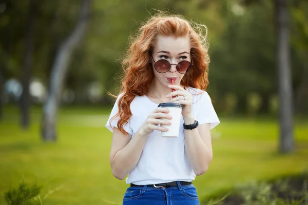 Hermosa chica pelirroja camina por el parque con una taza de café. La chica sonríe a la cámara. Una chica con gafas de sol y un reloj moderno en su brazo —  Fotos de Stock