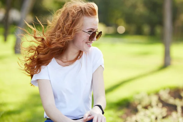 Retrato de una hermosa chica pelirroja que se sienta en el parque y mira hacia otro lado. El viento desarrolla su cabello y la chica sonríe —  Fotos de Stock