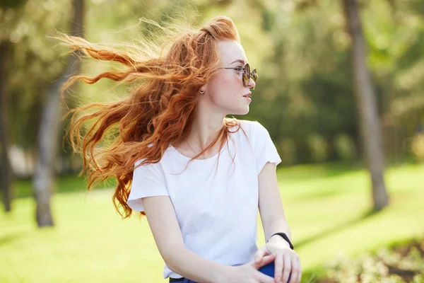 Retrato de una hermosa chica pelirroja que se sienta en el parque y mira hacia otro lado. El viento desarrolla su cabello y la chica sonríe —  Fotos de Stock