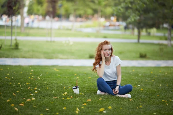Ένα όμορφο κορίτσι με κόκκινα μαλλιά κάθεται στο γκαζόν δίπλα σε ένα φλιτζάνι καφέ. Το κορίτσι κοιτάζει την κάμερα και χαμογελάει — Φωτογραφία Αρχείου