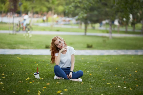Ένα όμορφο κορίτσι με κόκκινα μαλλιά κάθεται στο γκαζόν δίπλα σε ένα φλιτζάνι καφέ. Το κορίτσι κοιτάζει την κάμερα και χαμογελάει — Φωτογραφία Αρχείου