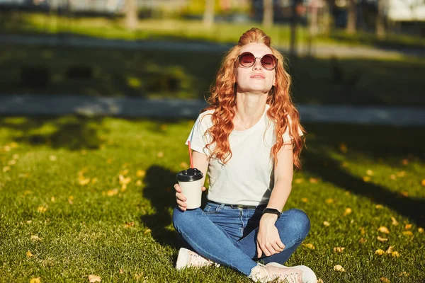 잔디밭에 앉아 아름 다운 빨간 머리 소녀입니다. 소녀의 손에 커피 한 잔. 젊은 아가씨는 아침 태양을 따뜻하게하고 그녀는 미소 — 스톡 사진