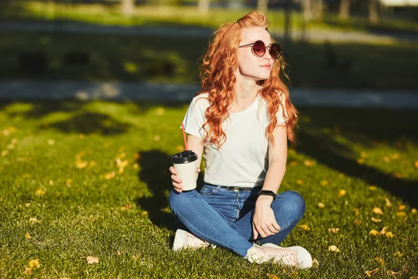 Çimlerin üzerinde oturan güzel kızıl saçlı kız. Kızın elinde bir bardak kahve. Genç bayan sabah güneşini ısıtıyor ve gülümsüyor. — Stok fotoğraf