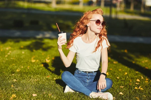Vacker rödhårig flicka sitter på gräsmattan. I händerna på flickan ett glas kaffe. Den unga damen värmer morgonsolen och hon ler — Stockfoto