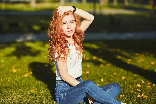 Una encantadora chica pelirroja, calentada por los rayos del sol, está sentada en un césped y posando para la cámara. La chica lleva una camiseta con jeans, gafas en la cara y un gadget moderno en el brazo. — Foto de Stock