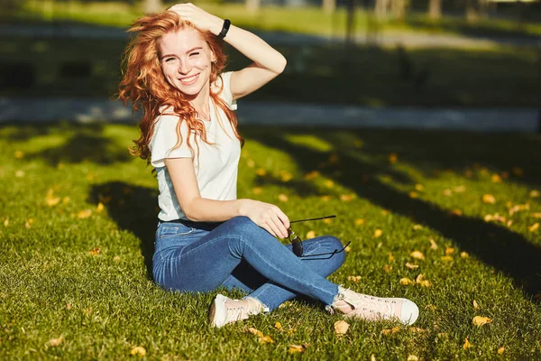 Güzel bir kızıl saçlı kız, güneş ışınları tarafından ısınmış, bir çim üzerinde oturuyor ve kamera için poz. Kız kot pantolon, yüzünde gözlük ve kolunda modern bir alet olan bir tişört giyiyor. — Stok fotoğraf