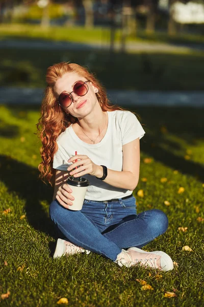 Vacker rödhårig flicka sitter på gräsmattan. I händerna på flickan ett glas kaffe. Den unga damen värmer morgonsolen och hon ler — Stockfoto