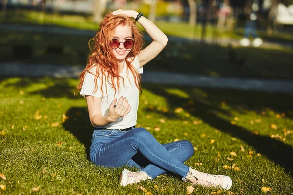 Une jolie fille aux cheveux roux, réchauffée par les rayons du soleil, est assise sur une pelouse et pose pour la caméra. La fille porte un T-shirt avec un jean, des lunettes sur son visage et un gadget moderne sur son bras — Photo